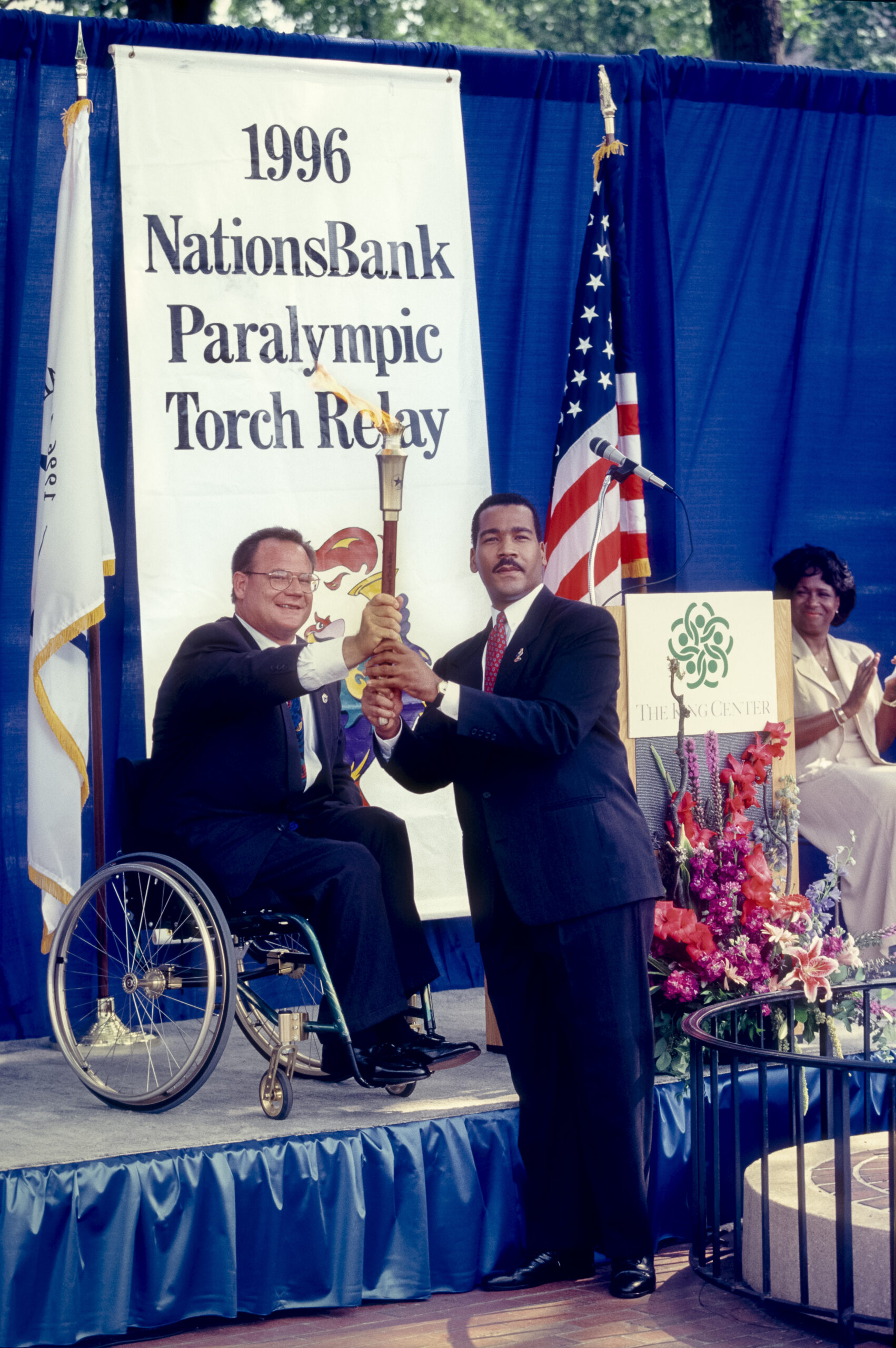 Two men holding a lit Paralympic torch while on a stage with the American flag in the background