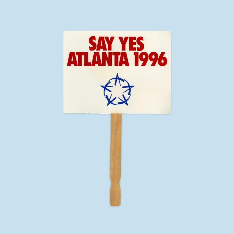 White handheld sign with red text that says Say Yes Atlanta 1996 and blue Olympic icon