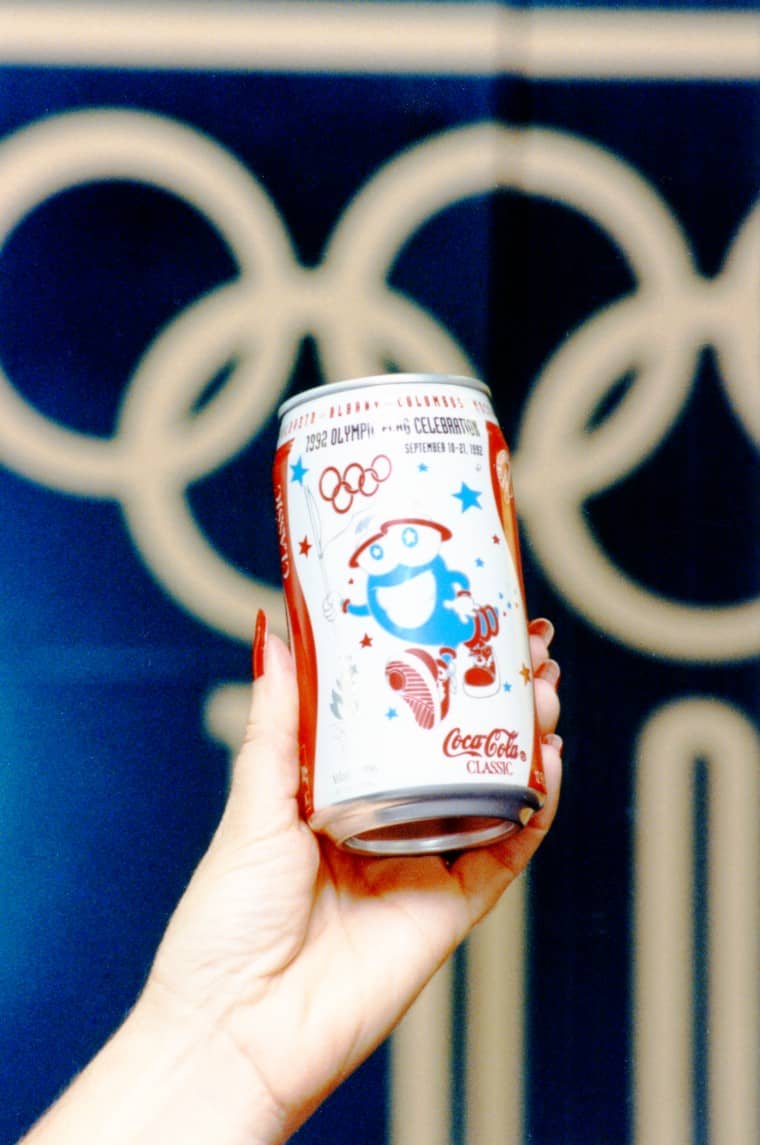 Hand holding a can of coke with Izzy mascot in front of an Olympic banner