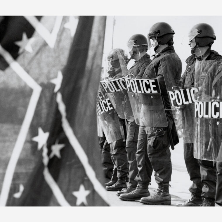 Members of law enforcement stand ready to protect Brotherhood March protestors from counter protestors including one in the foreground who is carrying a Confederate flag