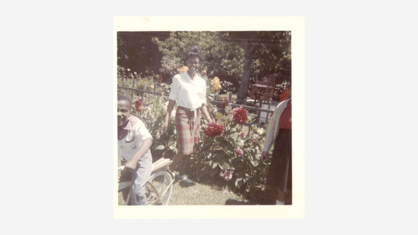 Two women with boy on bicycle in front of flower bed of dahlias and fence, circa 1960s